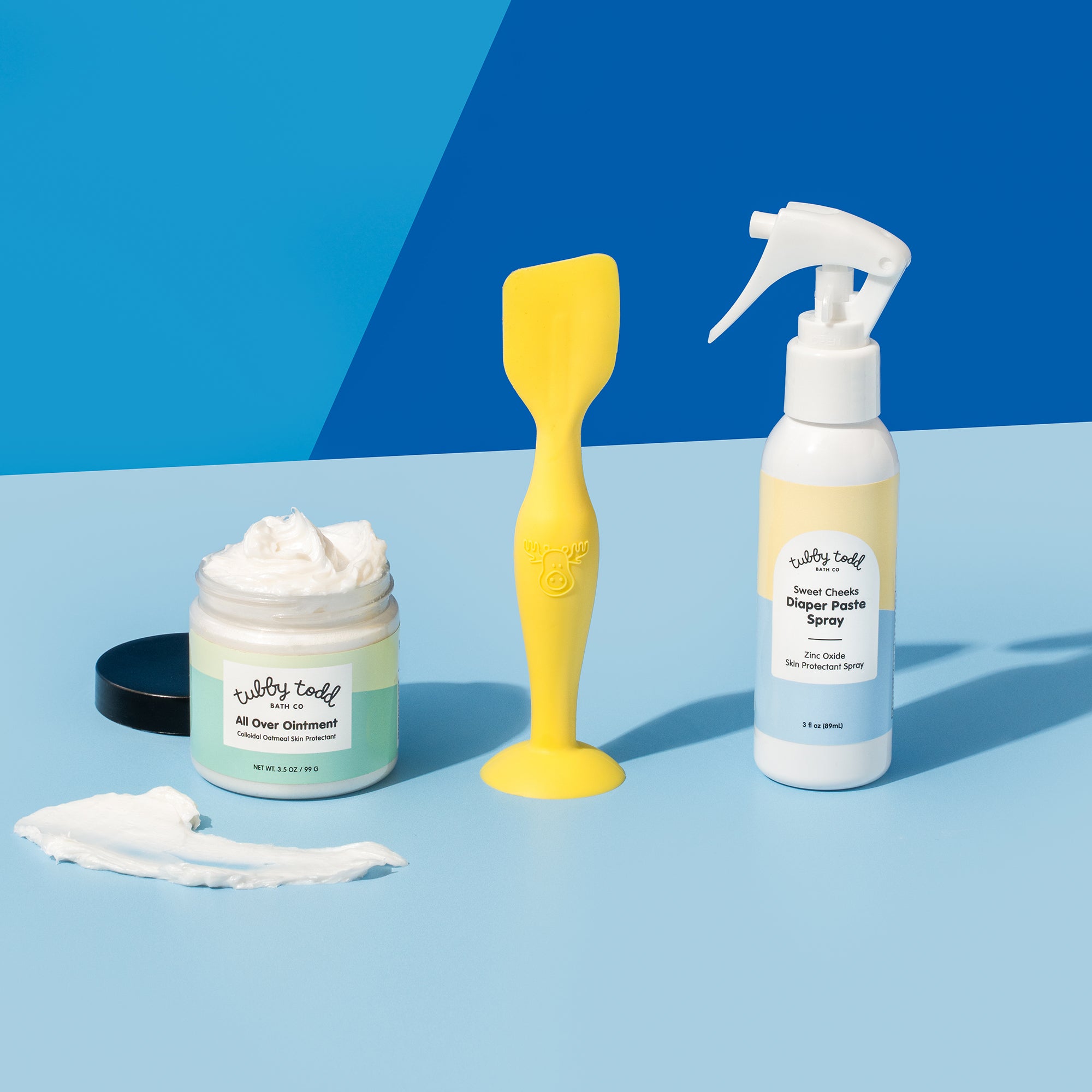 The 3.5 oz All Over Ointment, Silicone Spatula, and Diaper Paste Spray are standing on a blue background, and the texture of the AOO is showing next to the AOO jar.